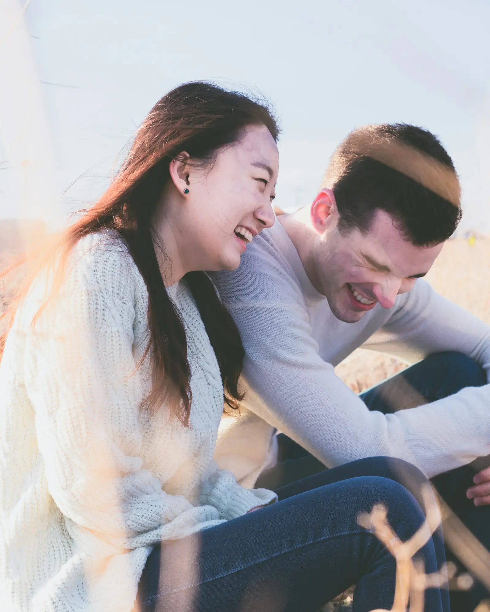A couple laughing together outdoors on a bright day, sitting close in the long grass